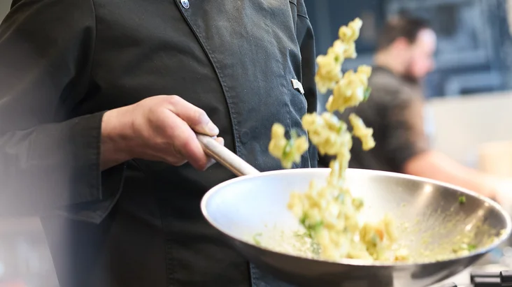 Der Koch steht in der Küche und bereitet Pasta in der Pfanne zu. 