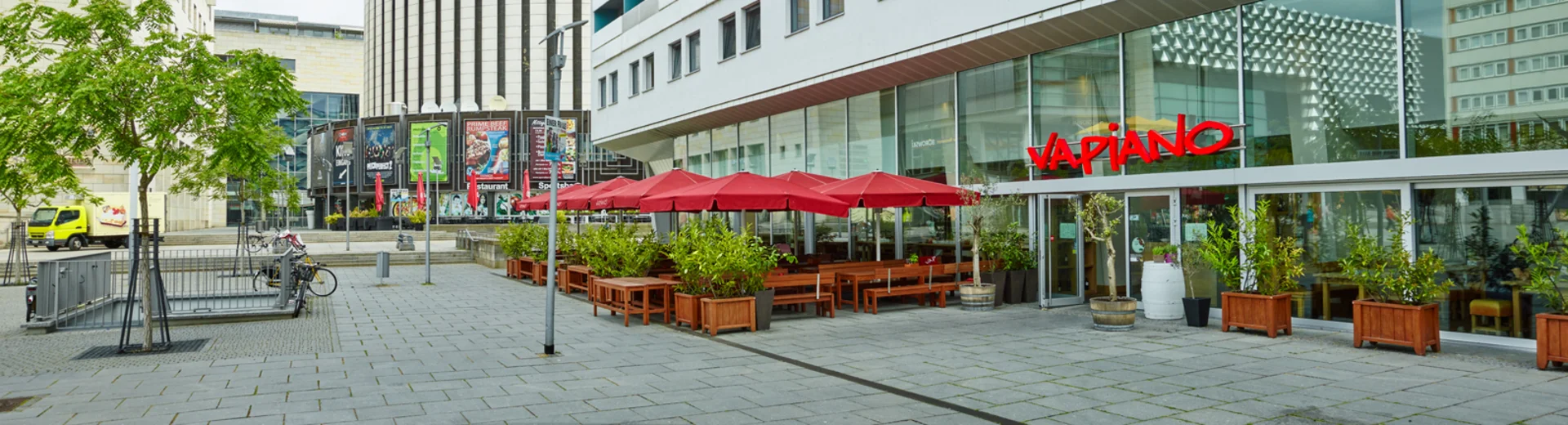 Außenansicht vom VAPIANO Dresden Prager Straße mit Bänken, Tischen und roten Sonnenschirmen im Außenbereich.