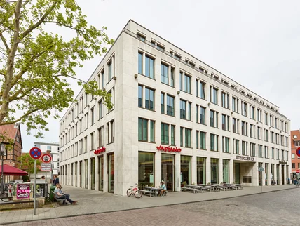 Modernes helles Eckgebäude mit großen Fenstern; Erdgeschoss Restaurant mit Außensitzplätzen an ruhiger Straßenkreuzung
