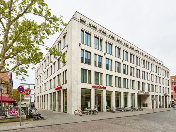 Modernes helles Eckgebäude mit großen Fenstern; Erdgeschoss Restaurant mit Außensitzplätzen an ruhiger Straßenkreuzung
