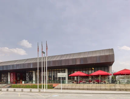 Flaches modernes Gebäude mit Glasfront und Metall­dach; davor Terrasse mit roten Schirmen und Fahnenmasten an Straße
