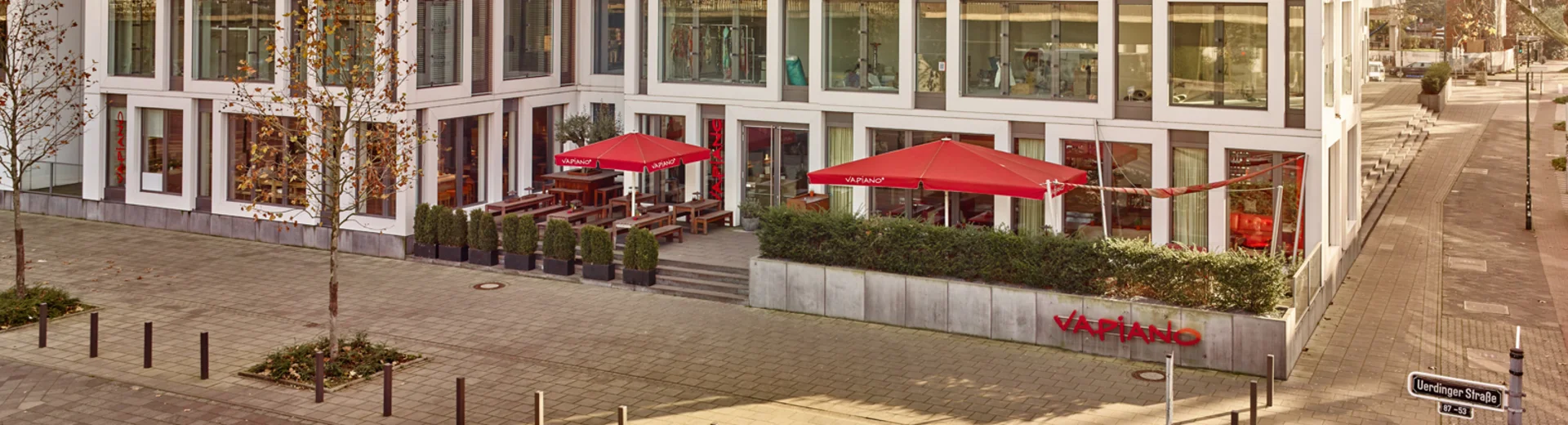Außenansicht vom VAPIANO Düsseldorf Golzheim mit Bänken, Tischen und roten Sonnenschirmen im Außenbereich. 