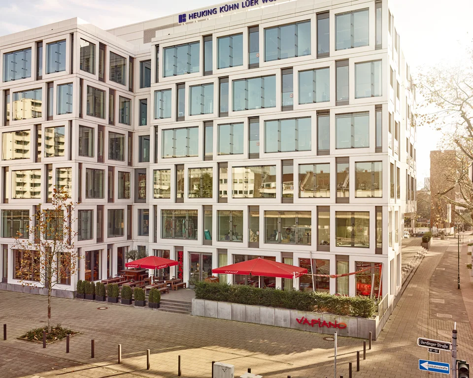 Außenansicht vom VAPIANO Düsseldorf Golzheim mit Bänken, Tischen und roten Sonnenschirmen im Außenbereich.