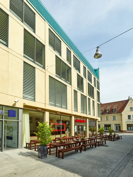 Modernes Gebäude mit heller Fassade und Lamellenfenstern; unten Restaurantterrasse mit Holzbänken und Pflanzen in ruhiger Straße

