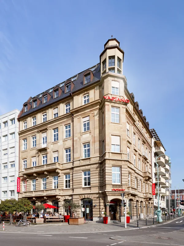Historisches Eckgebäude mit verzierter Sandsteinfassade; unten Restaurant mit roten Schirmen an belebter Straßenecke
