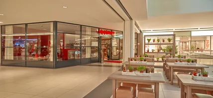 Modernes Einkaufszentrum innen; Restaurant mit Glasfront, Sitzbereich mit Tischen, Pflanzen und heller Beleuchtung