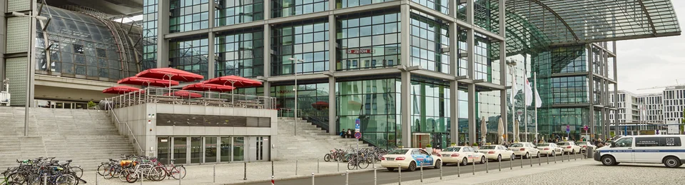Ein großes Gebäude am Hauptbahnhof Berlin mit roten Sonnenschirmen auf einer Terrasse vom VAPIANO Berlin Europaplatz.
