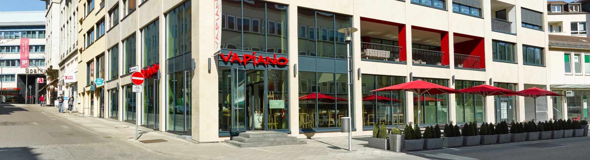 Außenansicht vom VAPIANO Kassel mit Bänken und roten Sonnenschirmen. 