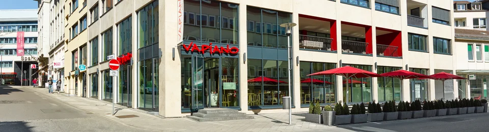 Außenansicht vom VAPIANO Kassel mit Bänken und roten Sonnenschirmen.