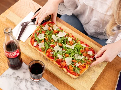 Eine Frau sitzt am Tisch mit einer belegten Pinsa und einem Getränk. 