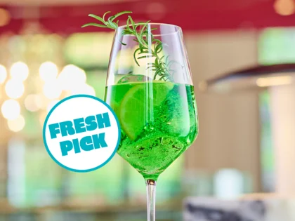 Ein leuchtend grüner Drink mit Limette und Rosmarin steht auf einer Bar, stilvoll in Szene gesetzt vor einem warm beleuchteten Hintergrund.