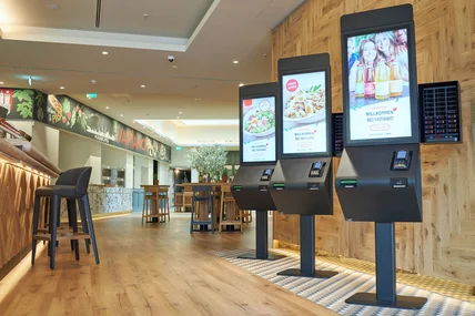 Modernes Restaurantinnere mit Holzdekor; drei Bestellterminals, Tresen, hohe Hocker und Tische im Hintergrund