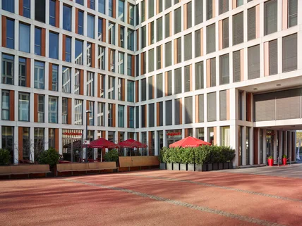 Großer moderner Gebäudekomplex mit Rasterfassade; Innenhof mit Sitzbänken, Pflanzen und Caféterrasse mit roten Schirmen
