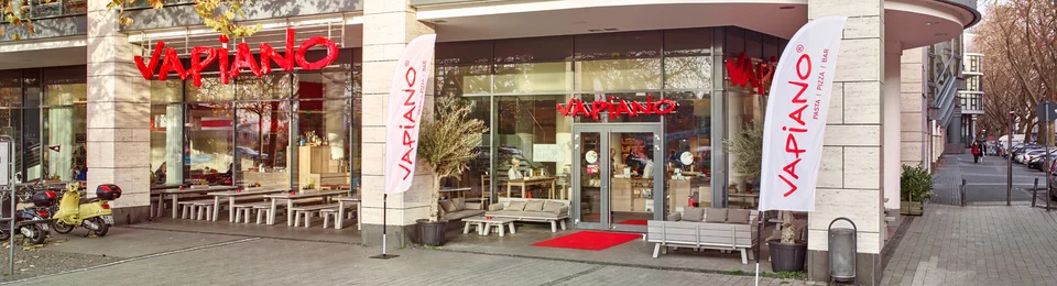 Außenansicht vom VAPIANO Köln Mediapark im Erdgeschoss eines mehrstöckigen Gebäudes mit Bänken in der Fußgängerzone. 