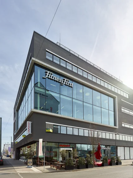 Modernes Eckgebäude mit großer Glasfassade; unten Restaurantterrasse mit Tischen, Pflanzenkübeln und roten Schirmen an Straße
