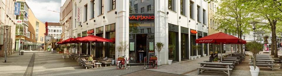 Außenansicht vom VAPIANO Hannover in der Fußgängerzone mit Bänken und roten Sonnenschirmen. 
