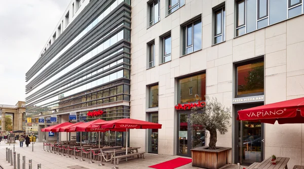 Modernes Gebäude mit Stein- und Glasfassade; Restaurantterrasse mit roten Schirmen, Holztischen und Pflanzkübel an Straße
