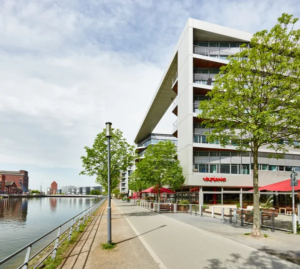 Modernes Gebäude am Fluss mit breitem Dachüberstand; Uferpromenade, Bäume und Restaurantterrasse mit roten Schirmen