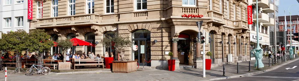 Außenansicht vom VAPIANO Mannheim im Erdgeschoss eines historischen Gebäudes mit Bänken und Sonnenschirmen. 