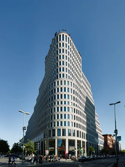 Keilförmiges Hochhaus mit vielen Fenstern an einer Straßenecke; unten Menschen und Autos, darüber klarer blauer Himmel
