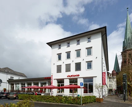 Schlichtes weißes Gebäude mit Satteldach; unten Restaurant mit Terrasse und roten Schirmen nahe Kirche und Parkplatz
