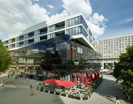 Modernes mehrstöckiges Gebäude mit Glasfassade und Auskragung; unten Restaurantterrasse mit roten Schirmen auf großem Platz
