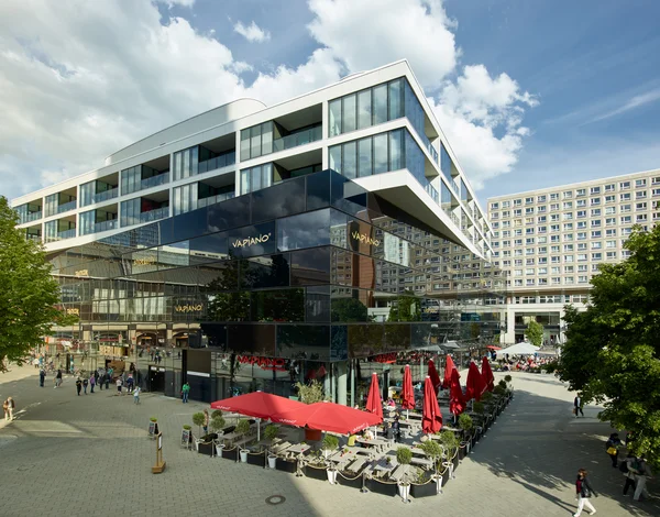 Modernes mehrstöckiges Gebäude mit Glasfassade und Auskragung; unten Restaurantterrasse mit roten Schirmen auf großem Platz

