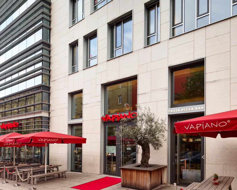 Außenansicht vom VAPIANO Stuttgart mit Bänken, Tischen und roten Sonnenschirmen in der Fußgängerzone. 