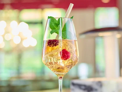 Ein pfirsichfarbener, spritziger Drink mit Beeren, Minze und Strohhalm steht auf einer Bar, stilvoll in Szene gesetzt vor einem warm beleuchteten Hintergrund.