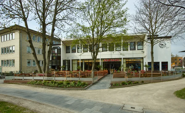 Flaches modernes Gebäude mit heller Fassade; große Außenterrasse mit Holztischen, Bäumen und breitem Gehweg davor
